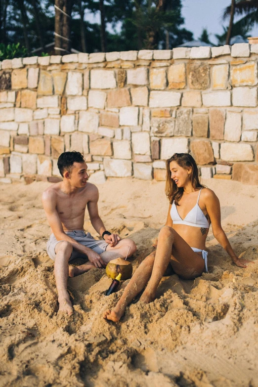 two people in bikinis sitting on sand next to wall