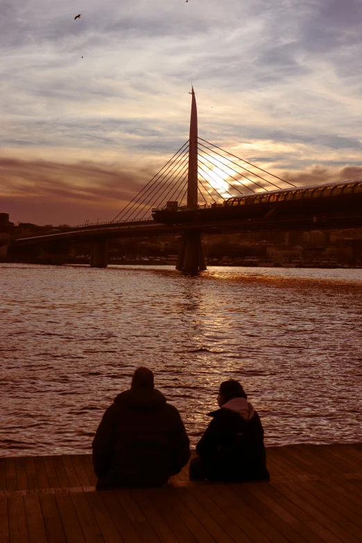 two people sit on a pier looking at the river