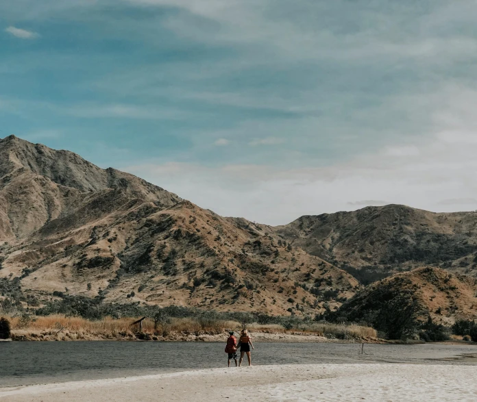 two people are walking together along the beach with mountains behind them