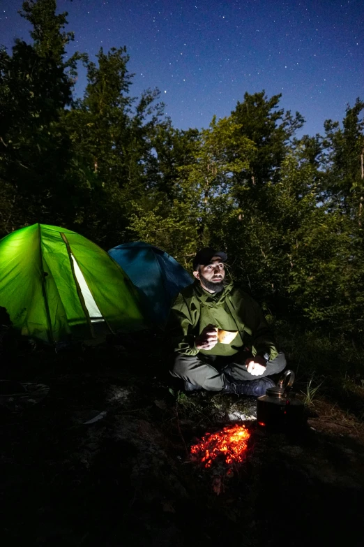 a person sits in the night beside a tent and campfire