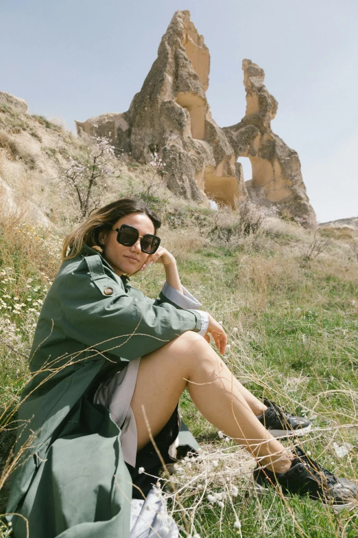 a woman in a coat and sunglasses sits in the grass by some big rocks