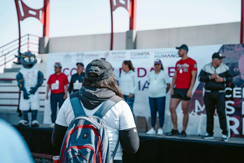 people watching an event with two men talking and the same woman holding a backpack