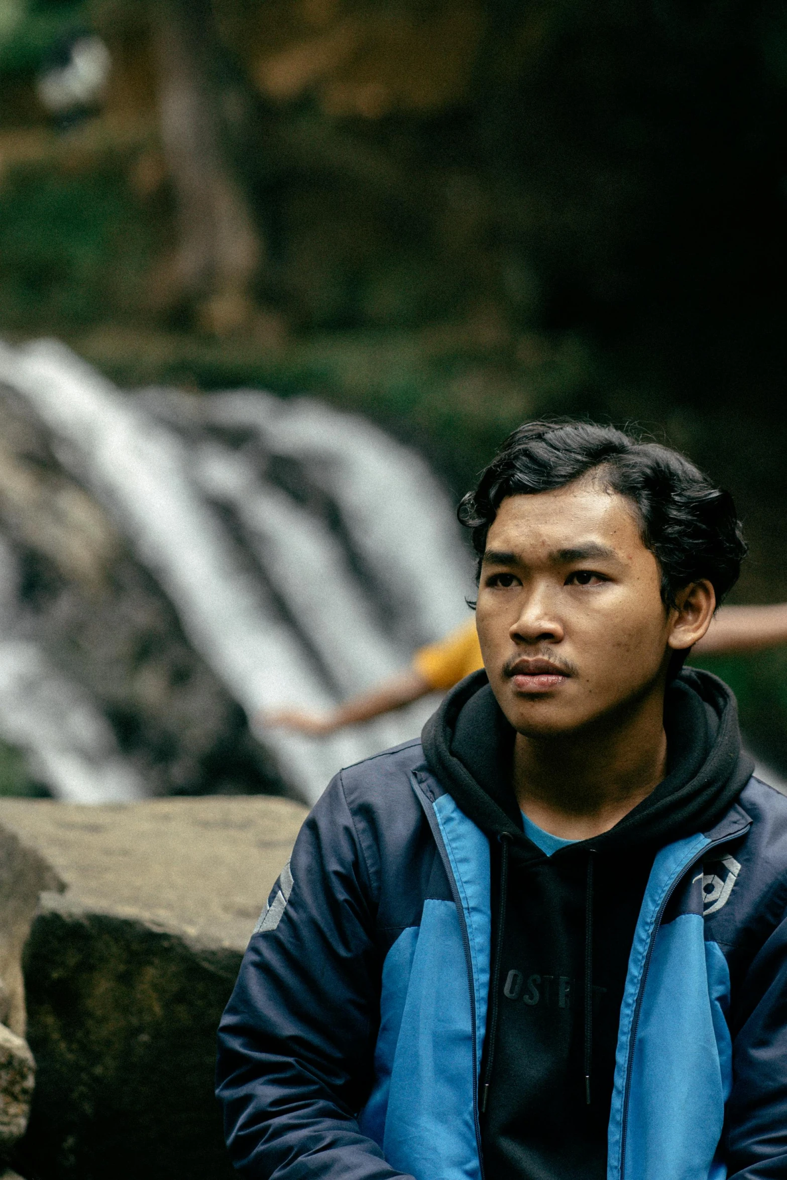 young man with hand on chin sitting near a waterfall