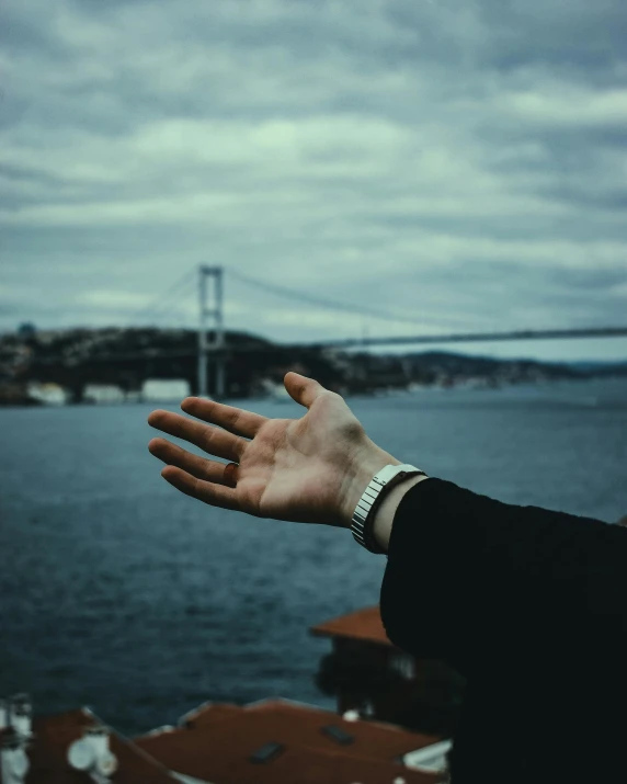 a man's hand reaching out towards the ocean and suspension bridge