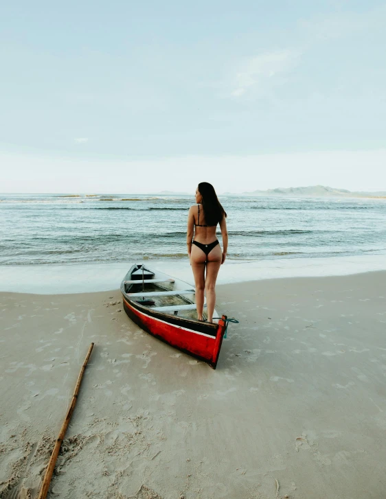 a woman in a bikini stands next to an empty boat on a beach