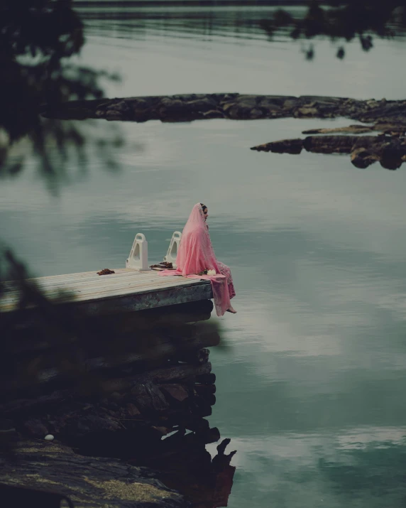 a person sitting on a dock in water with a pink dress on