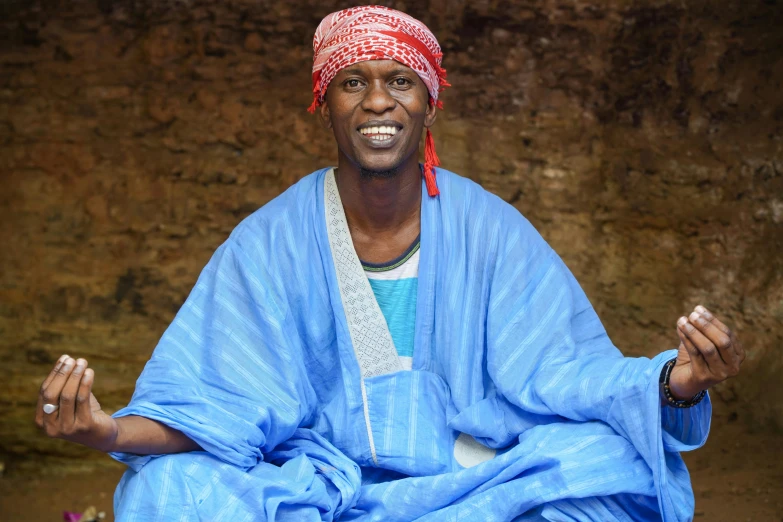 an african american man in blue robe and red headwrap sits on the ground smiling at the camera