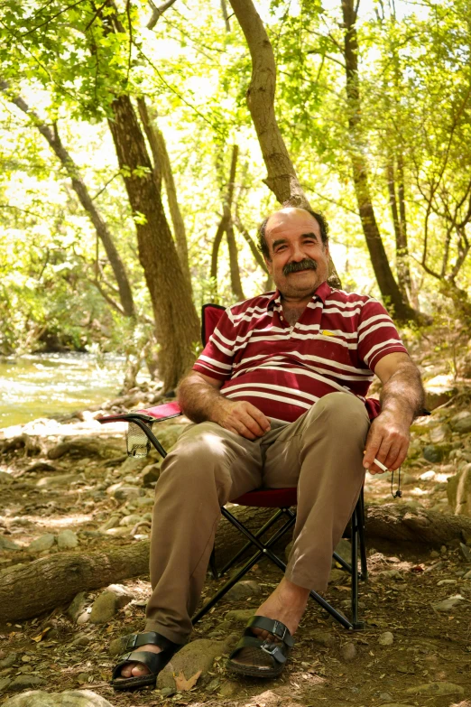 a smiling man sitting on a camp chair in the woods