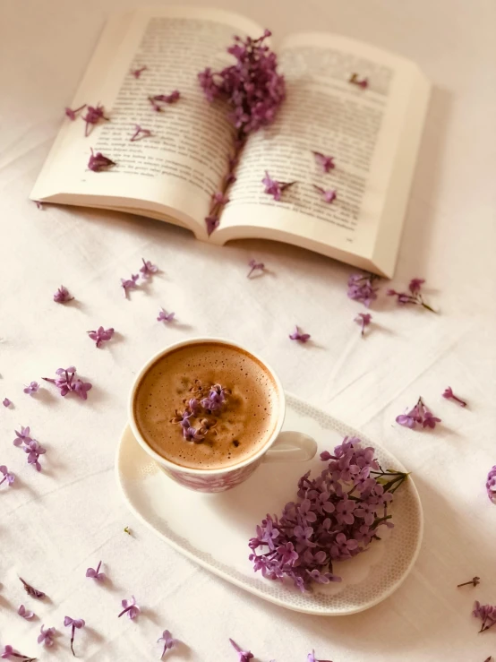 a coffee cup on a plate next to a book with purple flowers