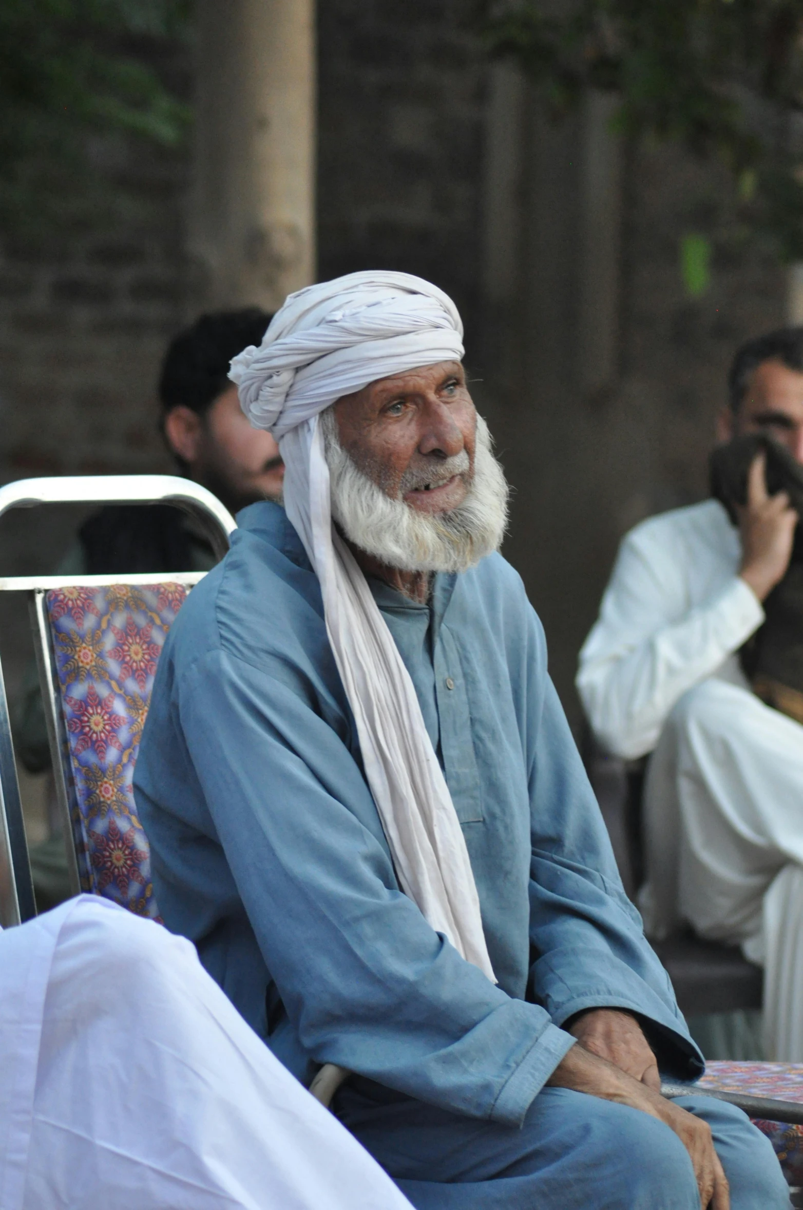 a man with long white hair and a white beard sits in a chair next to two other men wearing white clothing