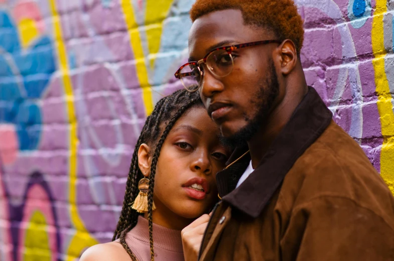 a man with glasses next to a woman standing against a colorful wall