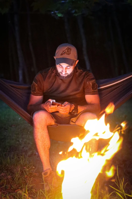 a man using his phone while sitting in a hammock by a fire