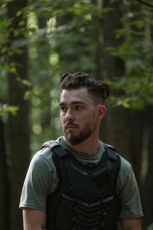 a man with a beard, wearing a black vest and standing in the woods