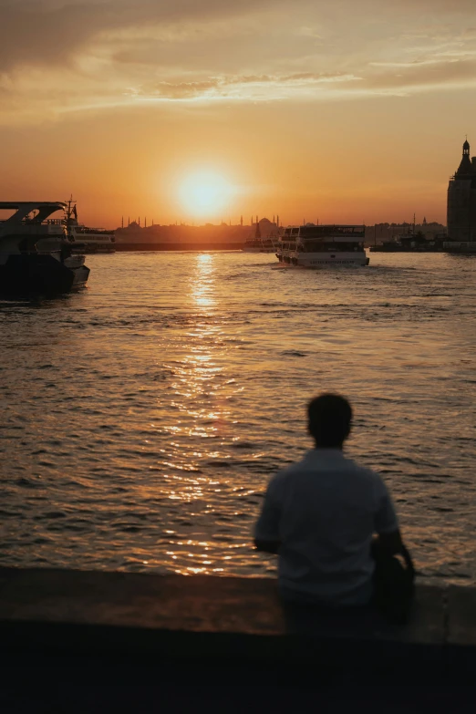 a man sits at the side of the water looking out at a boat