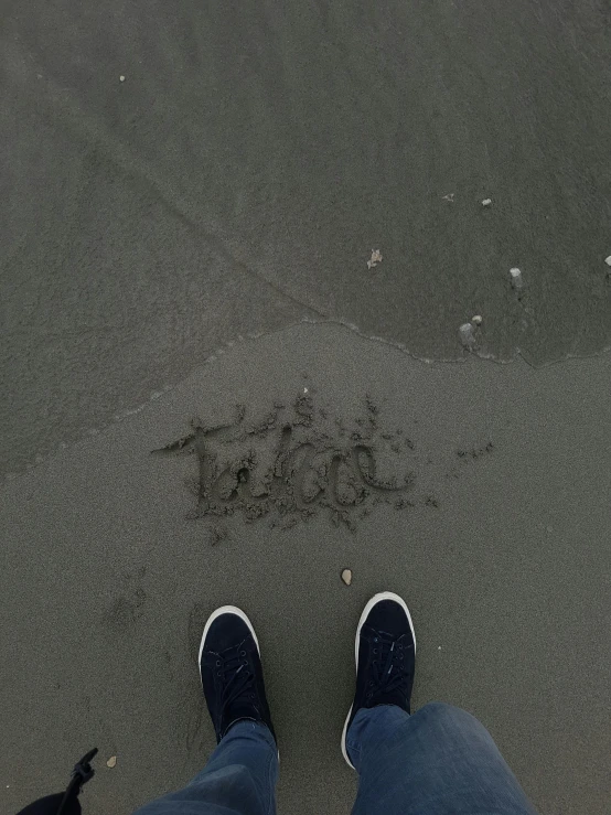 a person in blue jeans and black shoes stands on the beach with his feet up