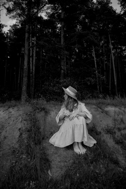 a woman wearing a dress and hat is sitting on the ground with trees