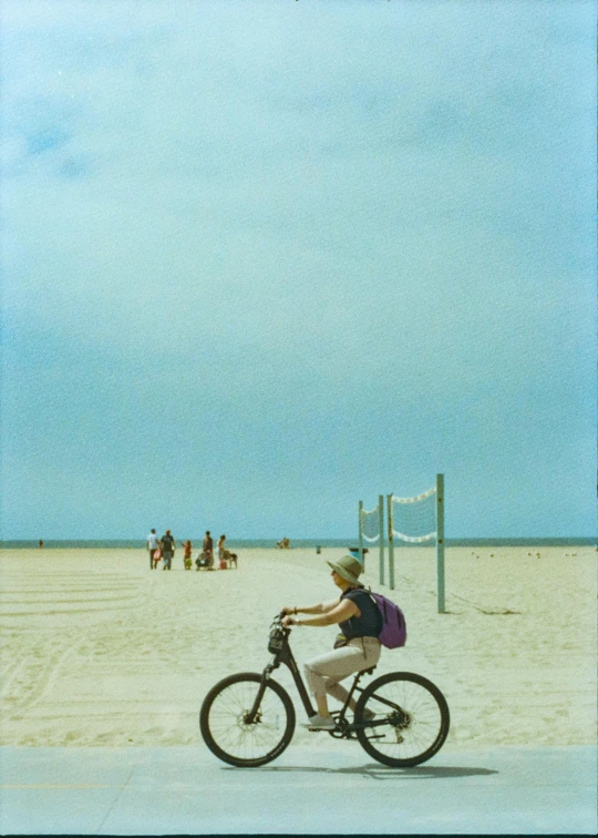 a man riding his bike down the beach with people playing in the background