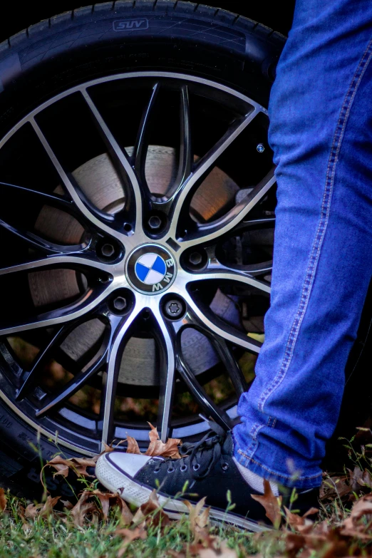 the black tire on a bmw car has been polished and a pair of blue jeans with worn down blue shoes is standing near it