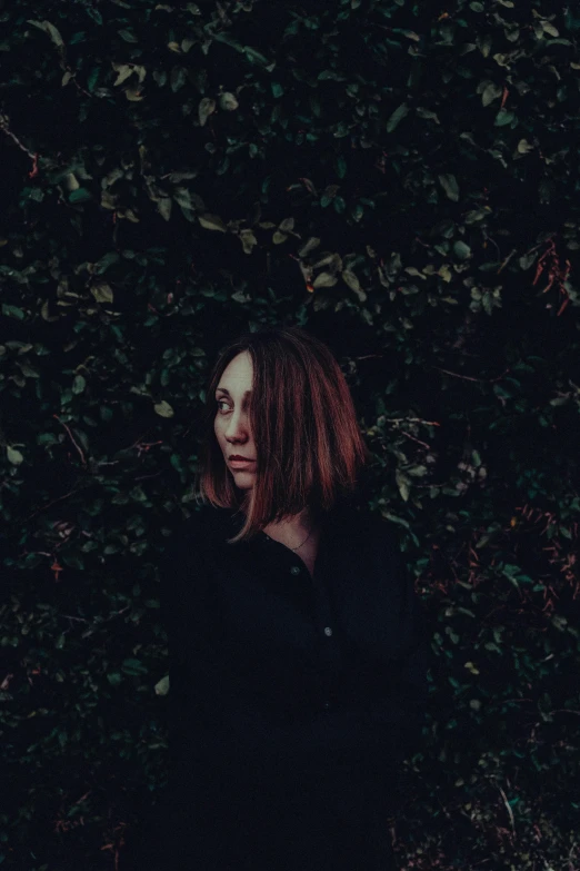 a woman is looking to the side while sitting outside in front of a bush