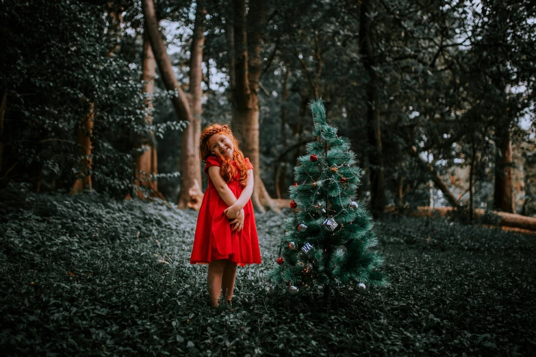 a girl in a red dress stands near a christmas tree