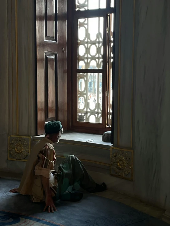 a little boy wearing a traditional islamic garb sits on the floor by an open window