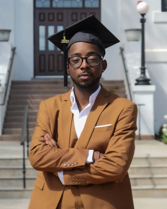 a man in graduation gown and cap standing with arms folded in front of stairs