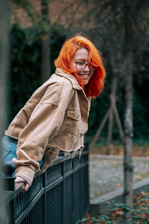 a redhead woman with glasses on leaning against a fence