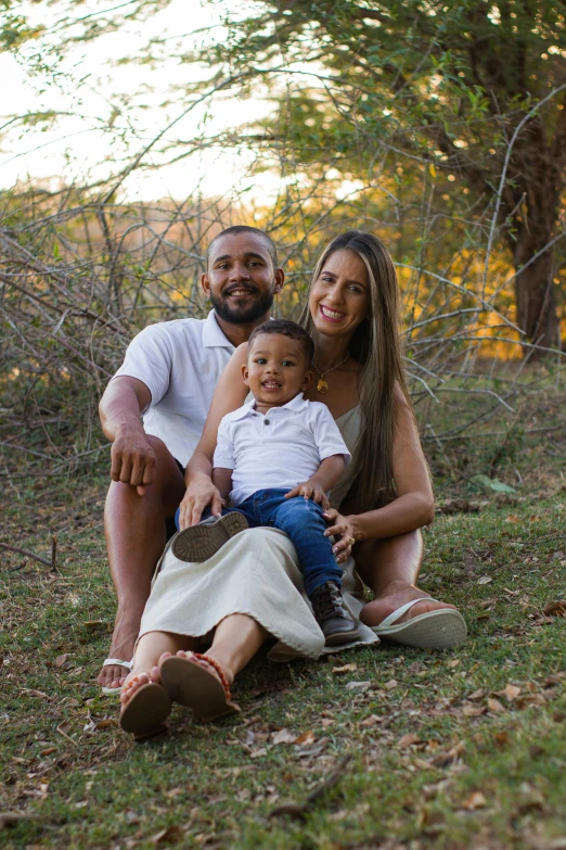 a man and woman with a baby sitting in the grass with a forest behind them