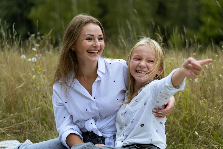 two girls are sitting in tall grass smiling and pointing