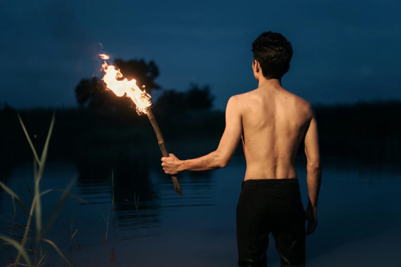 a shirtless man holding a burning torch stands near a body of water