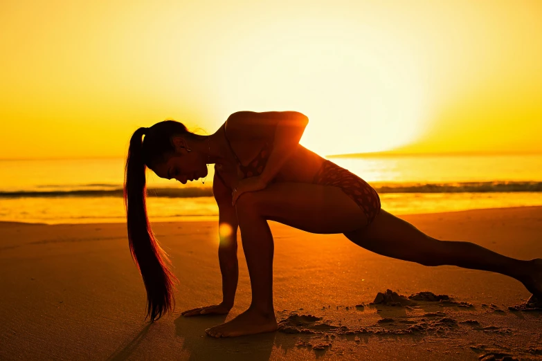 a woman stands on one leg, bending forward, in the sand and holding her hand up to her chin, as the sun sets overcast