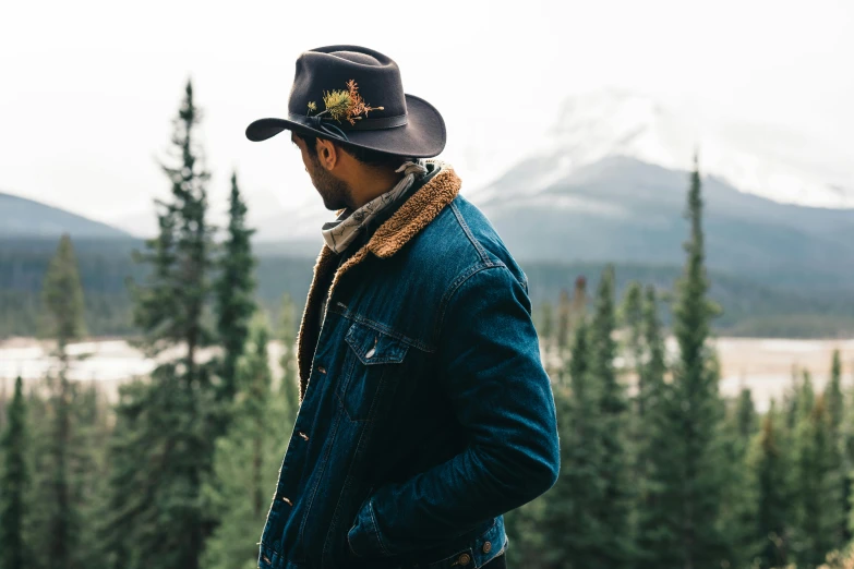 a man with a black hat, jeans jacket and a necklace looks out into the distance