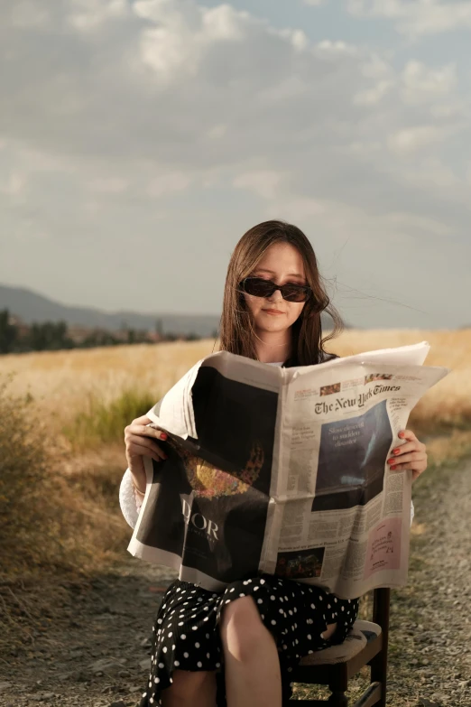 a woman in sunglasses is sitting on a bench and reading the newspaper