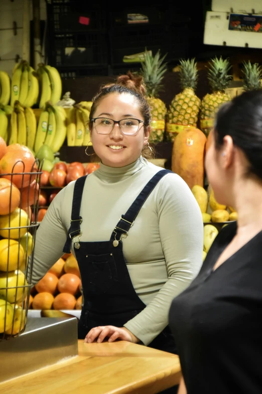 a woman smiles next to a stand with oranges, bananas and other fruit