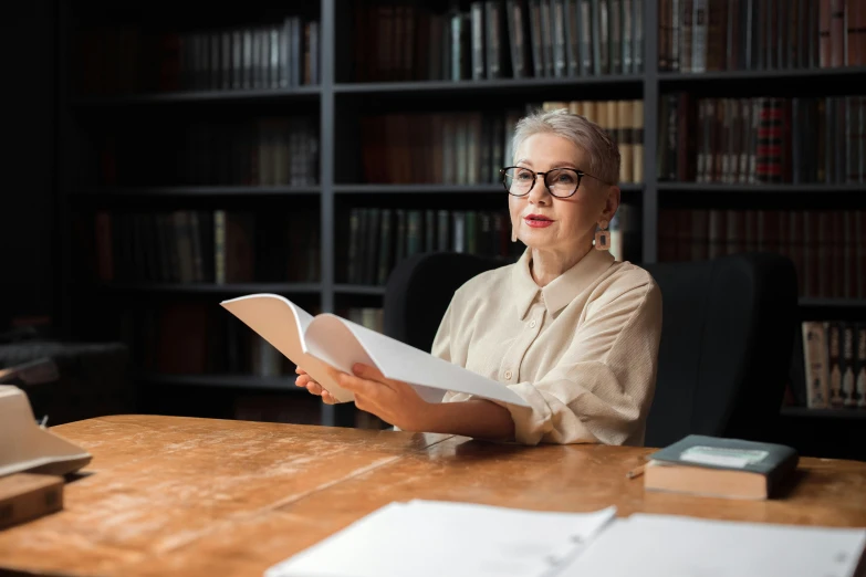 an older woman with glasses is reading a book in her office