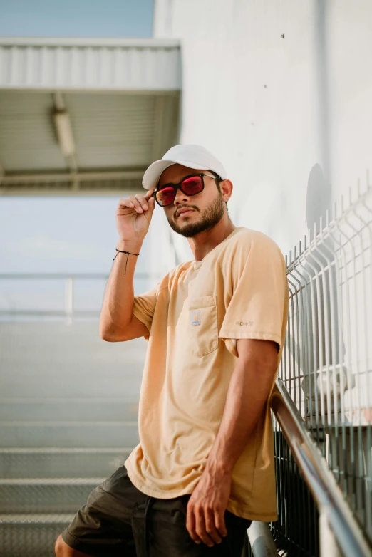 man wearing sunglasses and cap leaning against fence