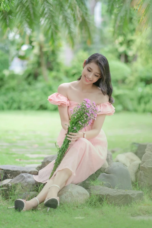 an asian woman in pink is sitting on the grass and holding a bouquet