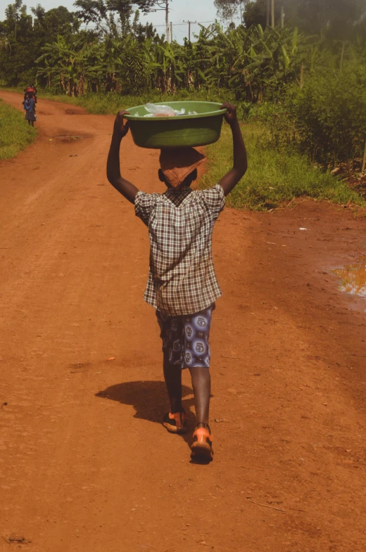 a boy walking down a dirt road carrying a green tray on his head