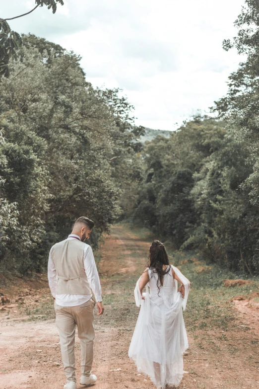 a bride and groom walking on a dirt road with lots of trees