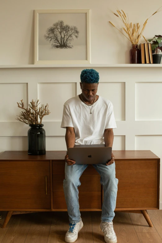 the man is sitting on the dresser and using his laptop