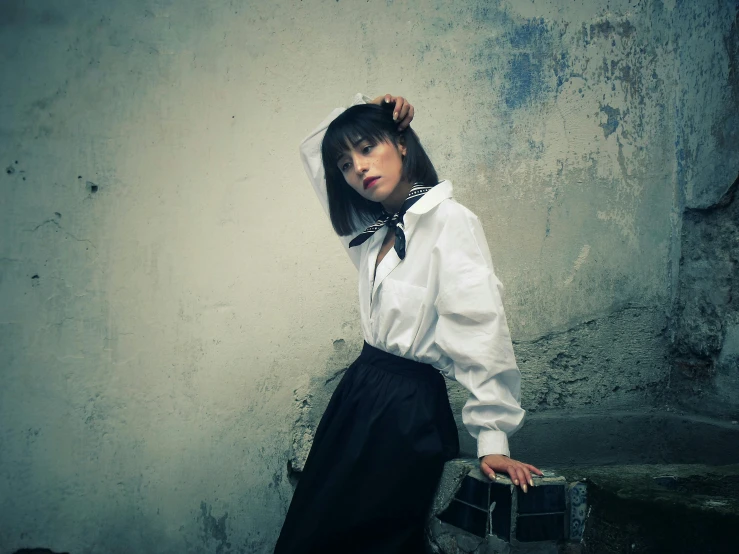 a woman wearing a white shirt and black skirt holds her head while standing in front of a dirty wall