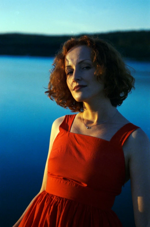 a woman in an orange dress stands with her hands on her hip looking towards the camera and a body of water in the background