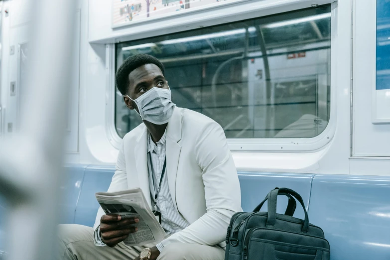 a man wearing a face mask sitting on a train reading a newspaper