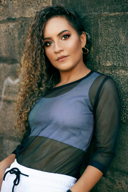 the young woman in mesh shirt is posing against a wall