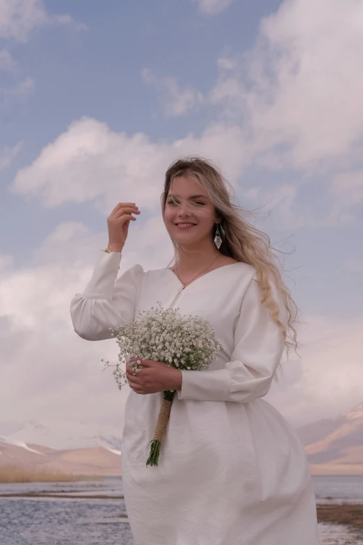 a woman in a white dress holding flowers by the water