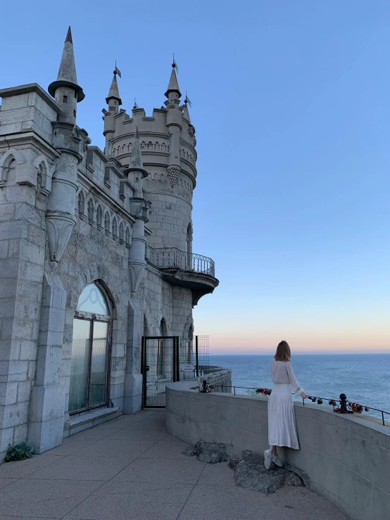 a woman stands on the wall next to a large castle by a blue ocean