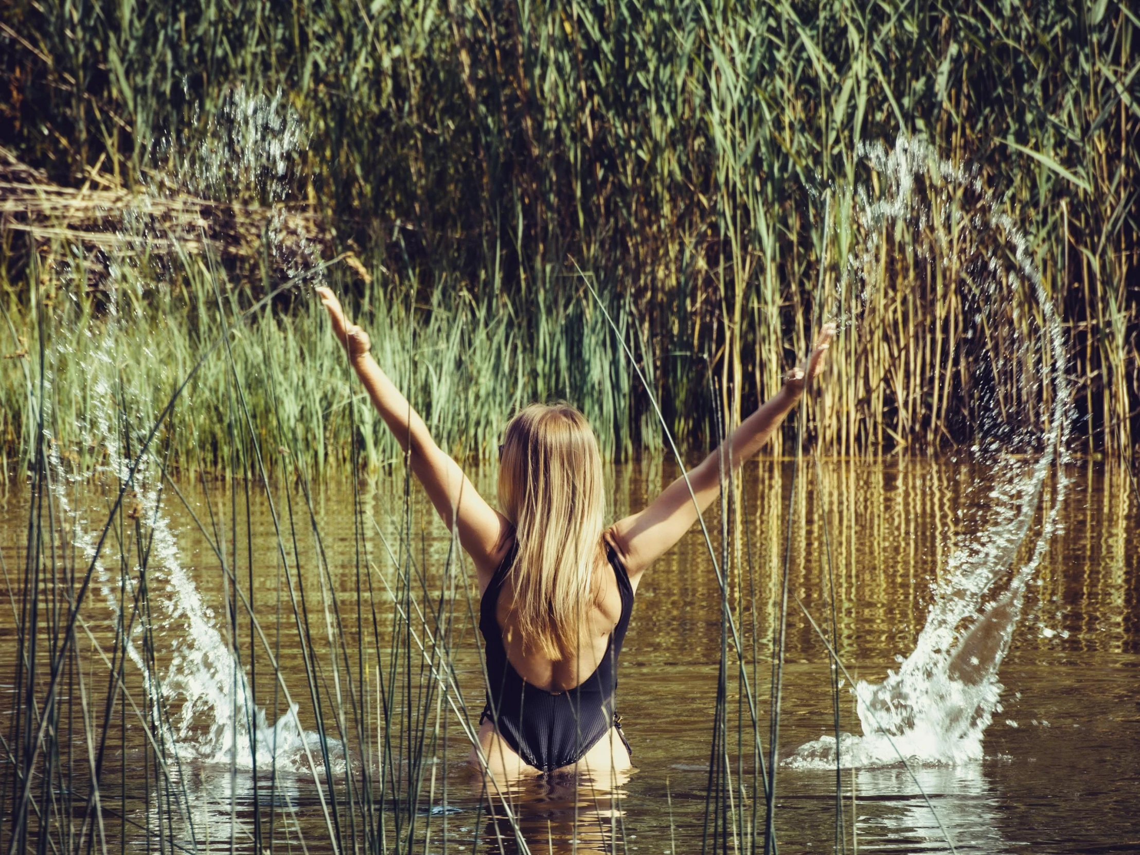 a girl wading in water holding her arms up as she is surrounded by water plants