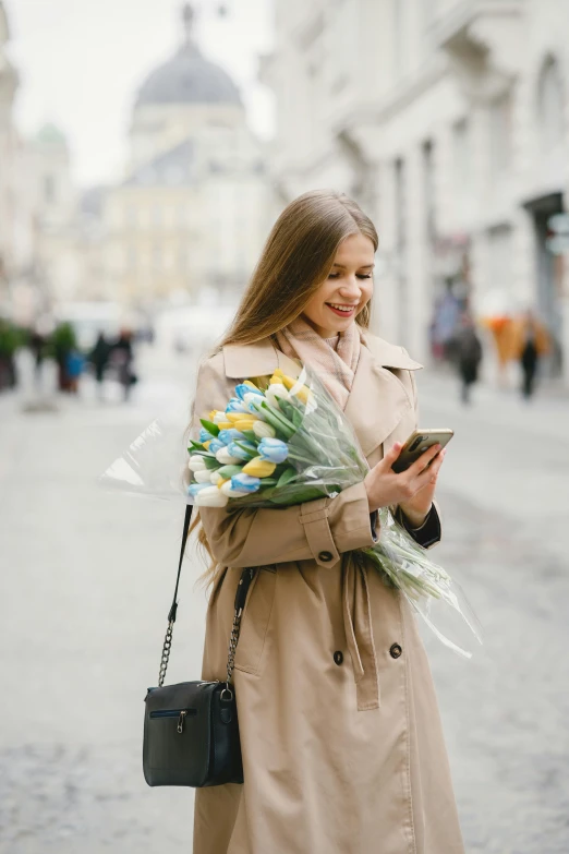 a beautiful young woman in trench coat holding a bouquet of flowers and a phone