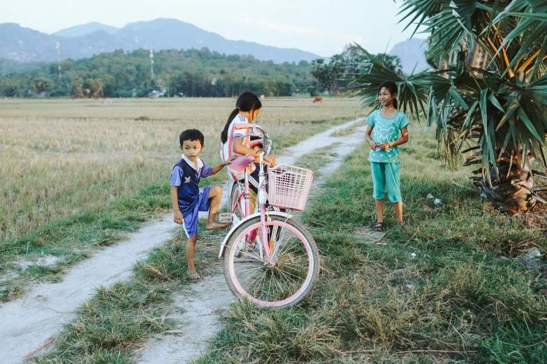 two children are standing next to a pink bicycle on the side of a dirt road