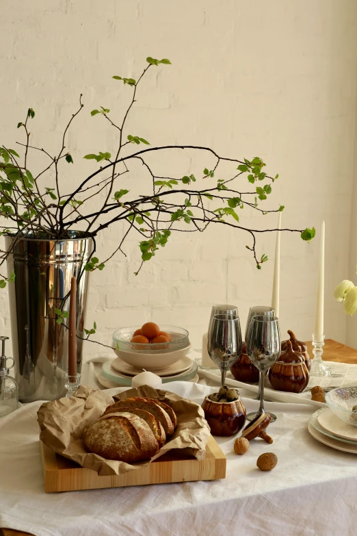 a table with a vase of green leaves, bread and fruit on it